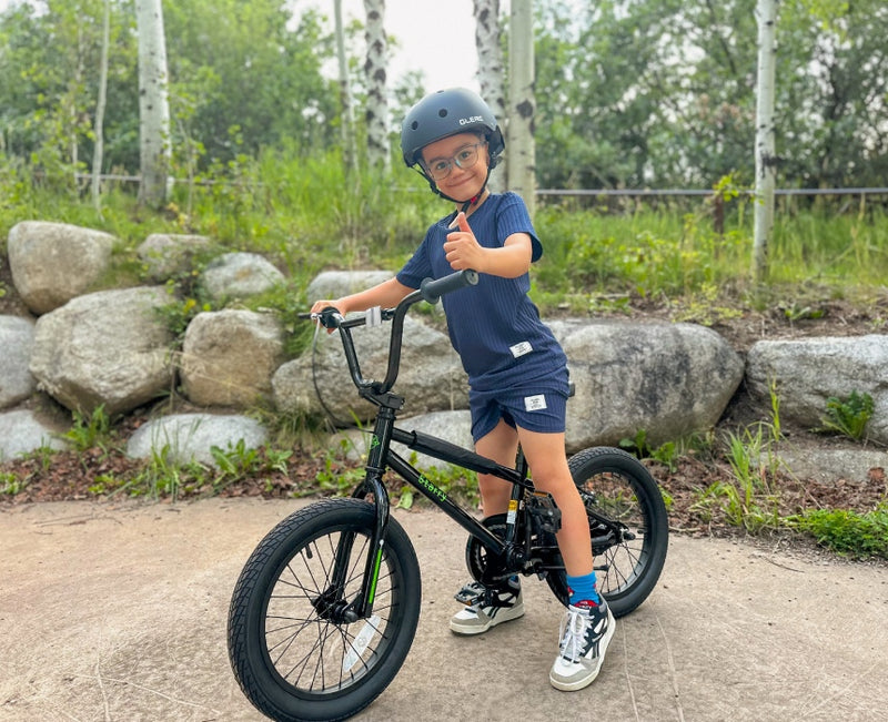 Boy in a black helmet giving a thumbs-up while sitting on a black kids' BMX bike on a stone path