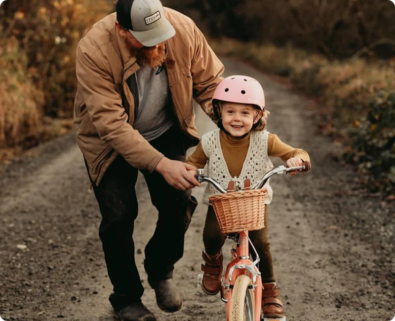 Father helping smiling daughter in a pink helmet ride a coral kids' bike with basket on a country trail