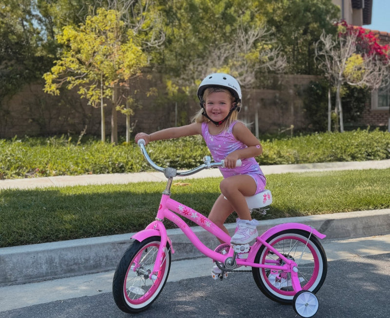 Smiling girl in a white helmet riding a pink kids' bike with training wheels on a sunny suburban street