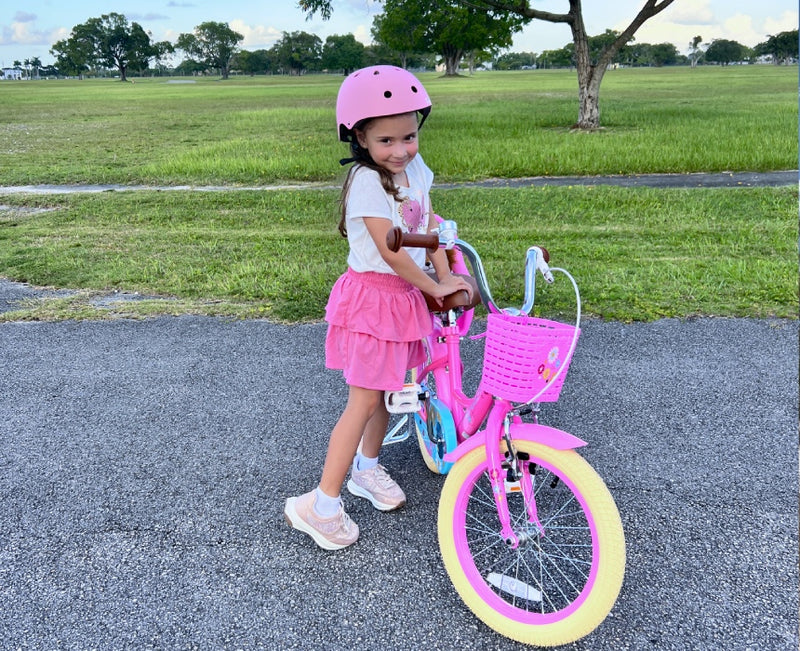 Girl in a pink helmet standing with a pink kids' bike featuring a flower basket on a park pathway
