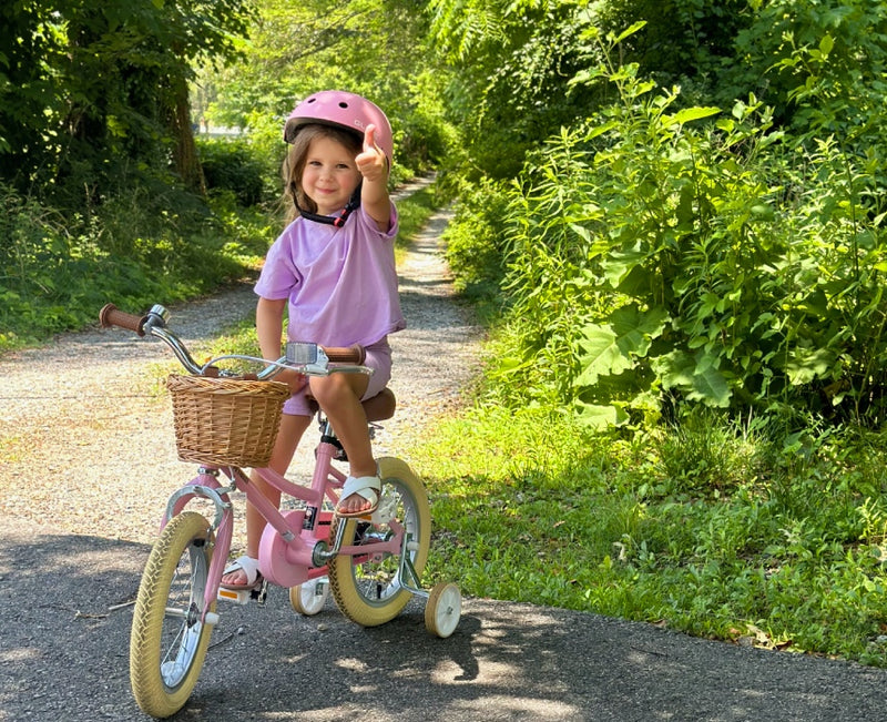 Girl in a pink helmet giving thumbs-up on a pink kids' bike with wicker basket and training wheels on a trail