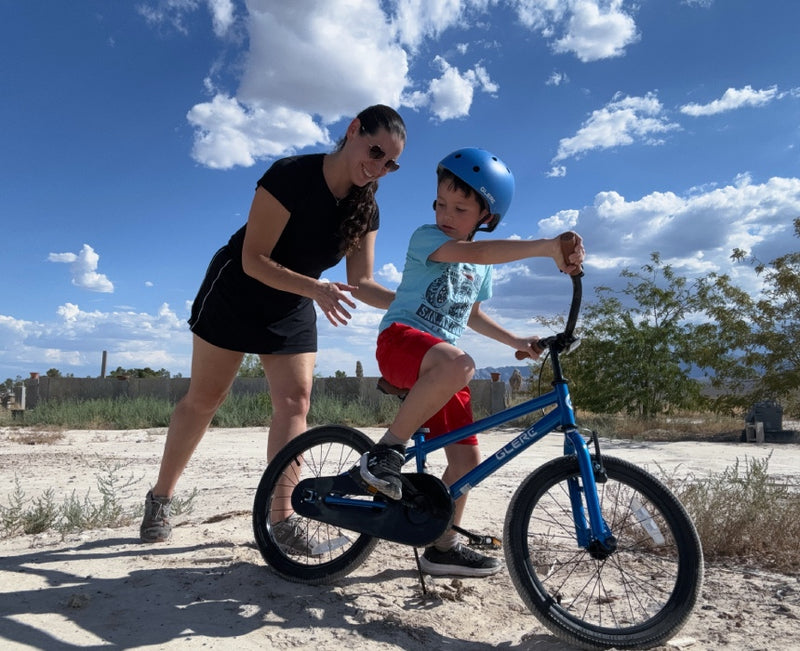 Mother helping her son in a blue helmet learn to ride a blue kids' BMX bike outdoors under a sunny sky