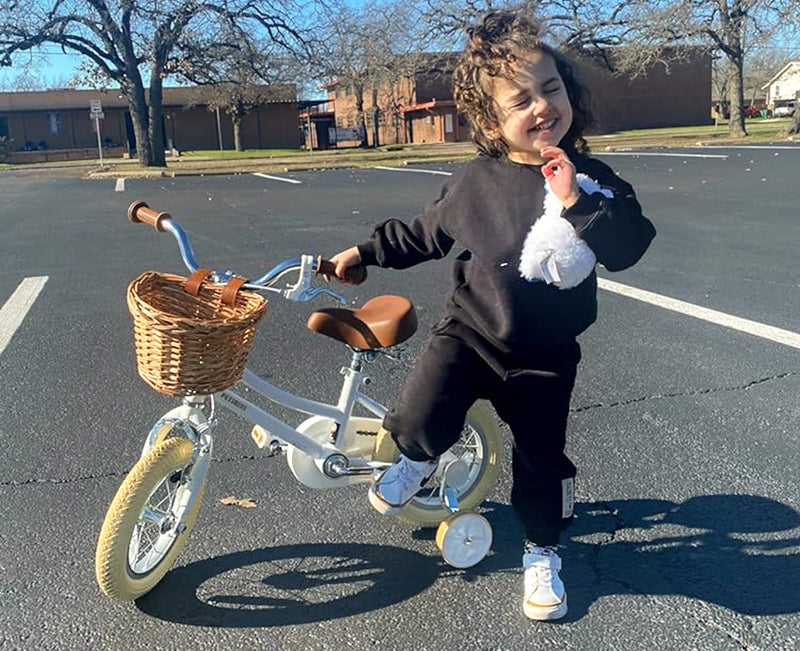 Laughing toddler holding a plush toy beside a white classic kids' bike with wicker basket and training wheels